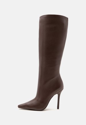 Botte haute en cuir brun foncé à talon haut avec un bout pointu et un talon aiguille fin, montrée sur un fond clair uni.