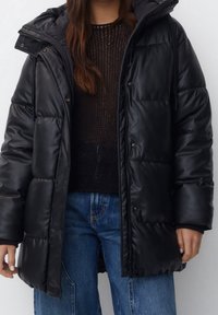 Doudoune noire à finition brillante, dotée d'une capuche, de boutons-pression et d'une coupe décontractée. Portée par-dessus un pull en maille foncé et un jean bleu.