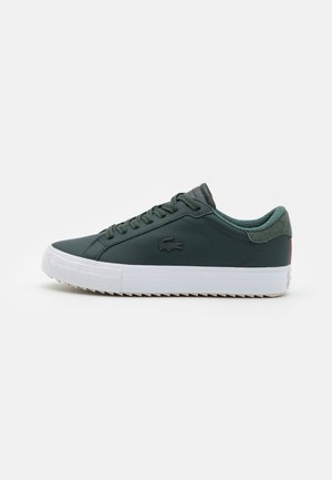 Zapatilla deportiva baja de color verde oscuro con suela blanca, cordones y un pequeño logo de cocodrilo en el lateral.