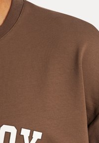 Sudadera marrón con textura acanalada, cuello redondo y detalles en texto blanco en la parte delantera. Diseño de tela suave.