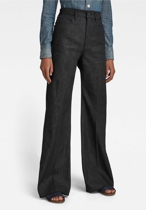 Personne portant un pantalon noir taille haute à jambes larges avec un bouton et une fermeture éclair à l'avant, associé à une chemise en denim bleu et des sandales à bouts ouverts.