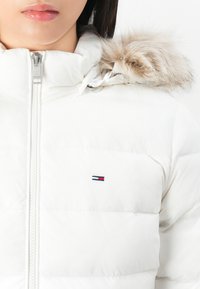 Frau trägt eine weiße Steppjacke mit pelzbesetzter Kapuze, zugezippt, mit einem kleinen gestickten Flaggenlogo auf der Brust.