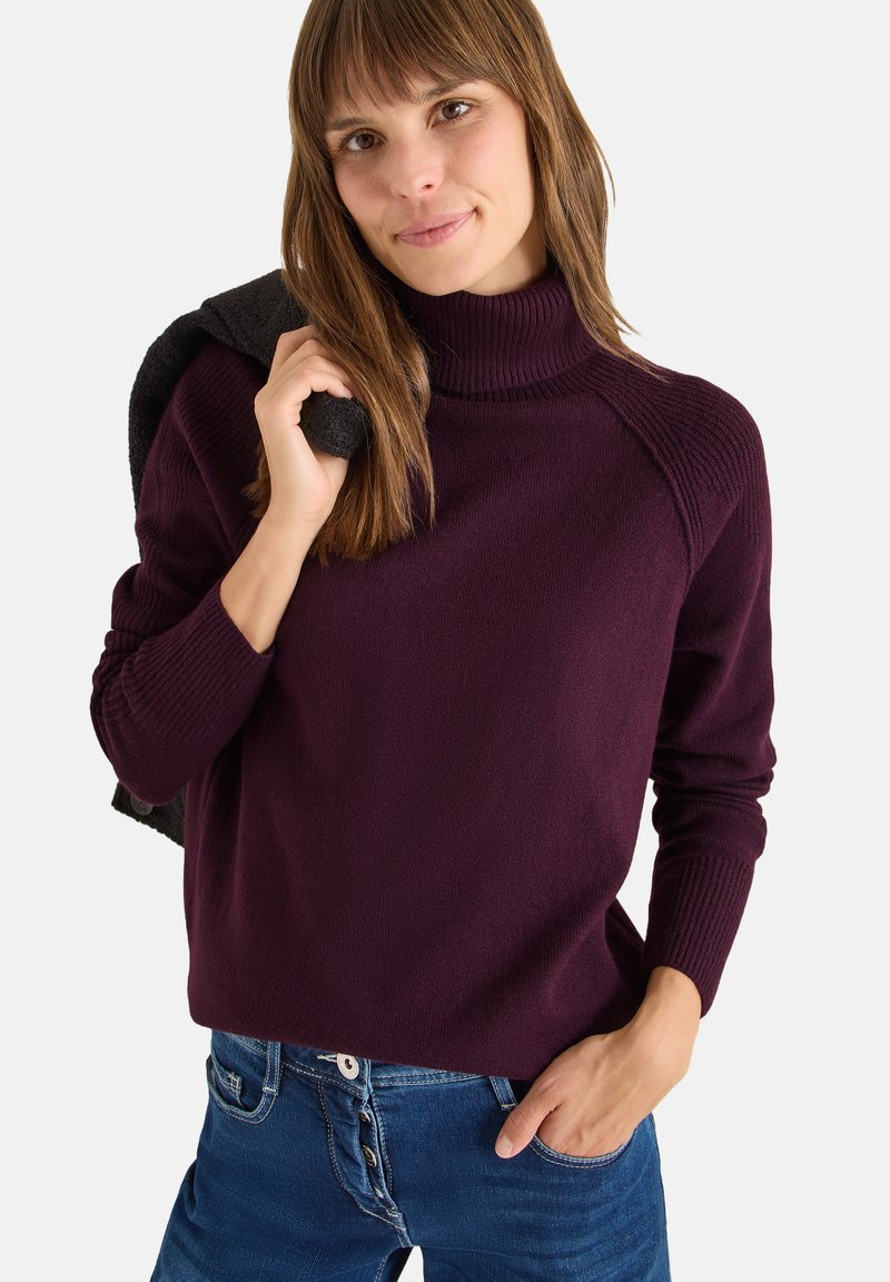 Maglione a collo alto color merlot con dettagli a coste, texture morbida e vestibilità rilassata, abbinato a jeans blu. Maglione scuro appoggiato sulla spalla.
