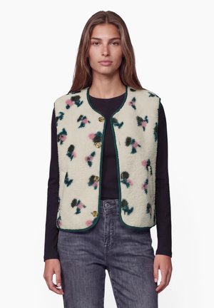 Femme portant une veste polaire crème avec des motifs floraux verts et roses sur un t-shirt noir à manches longues et un jean gris.