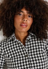 Chemise à motif pied de poule noir et blanc avec un col, deux boutons et un tissu texturé. Une peau lisse et un collier simple complètent le look.