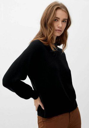 Young woman with wavy light brown hair wearing a black long-sleeve sweater and brown pants, standing with one hand on her hip.
