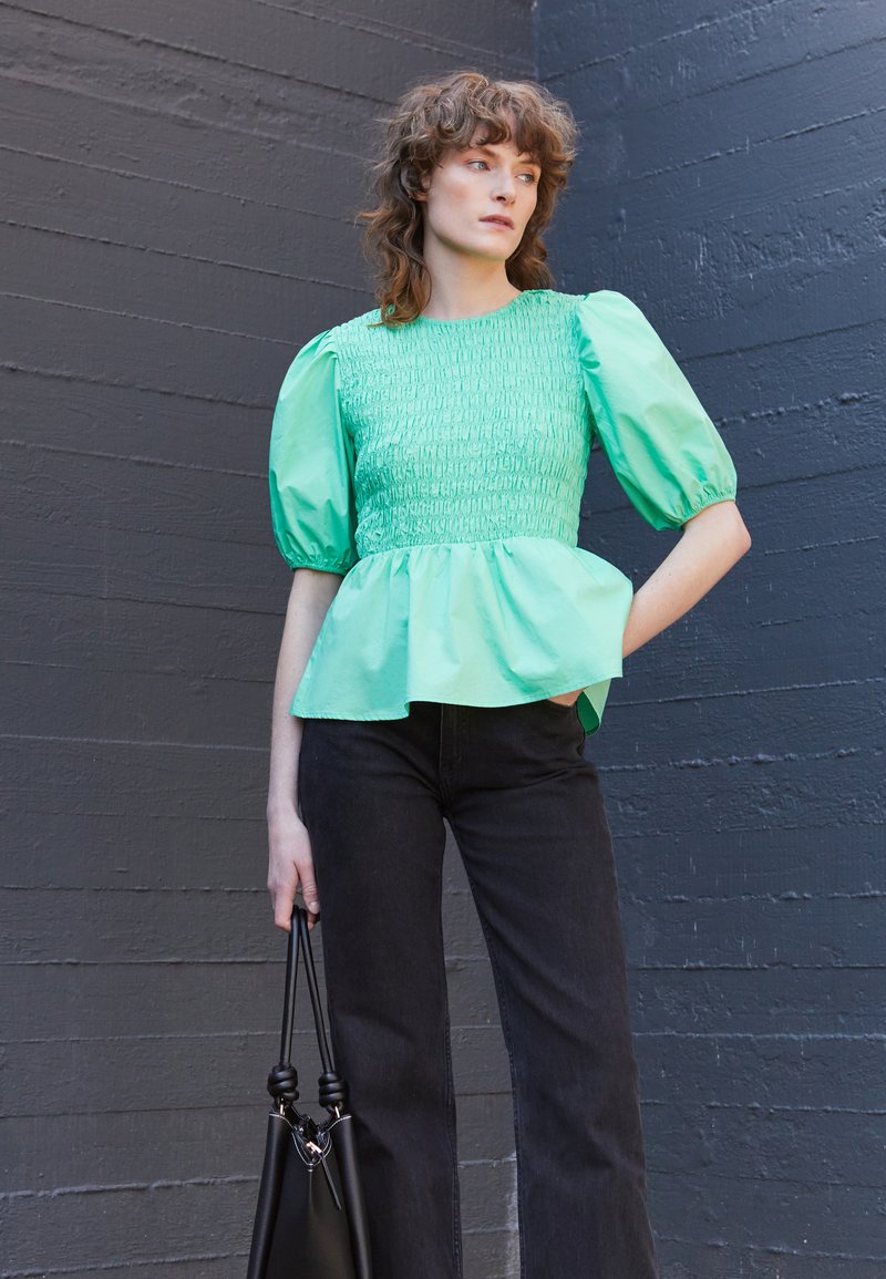 Top peplum verde-menta com corpete franzido e mangas bufantes, combinado com jeans pretos de perna larga e uma mala preta.