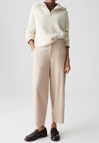 Personne portant un pull côtelé crème avec demi-fermeture éclair, pantalon large beige et mocassins en cuir marron foncé, debout avec une main dans la poche.