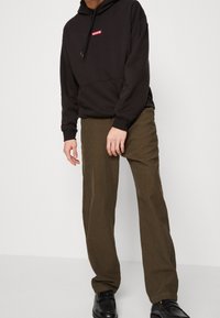 Zwarte hoodie met rood "Levi's" logo, gecombineerd met bruine broek met ruitpatroon. Model draagt zwarte schoenen en staat voor een effen achtergrond.