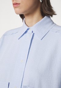 Jasnoniebieska koszula z kołnierzykiem wykonana z teksturowanej tkaniny. Posiada białe guziki oraz prosty krój z detalem kieszonki z przodu.