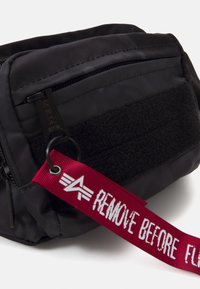 Borsa in nylon nera con chiusura a zip, caratterizzata da una toppa in velcro nera texturizzata e una targhetta di tessuto rossa con la scritta "REMOVE BEFORE FLIGHT".