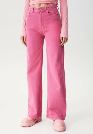 Personne portant un jean rose taille haute à jambes larges avec poches avant et des chaussures roses à semelle épaisse, associée à un crop top rose à manches longues.