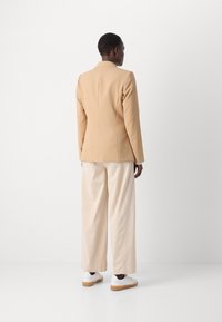 Blazer beige avec un design structuré, associé à un pantalon ample de couleur crème. Des baskets blanches avec une semelle en caoutchouc complètent la tenue.
