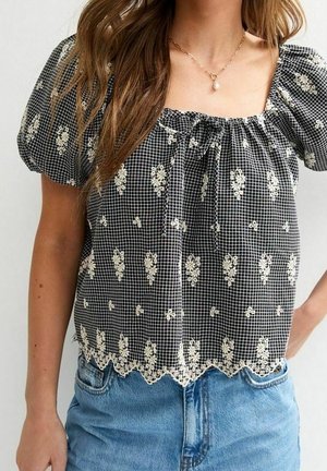 Kvinde iført en sort bluse med puffede ærmer, hvid blomsterbroderi og bølget kant, sat sammen med blå jeans.