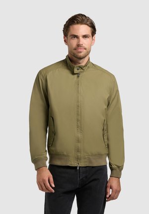 Mann trägt eine olivgrüne Jacke mit Reißverschluss, Knopfkragen und Seitentaschen, steht vor einem schlichten hellgrauen Hintergrund.