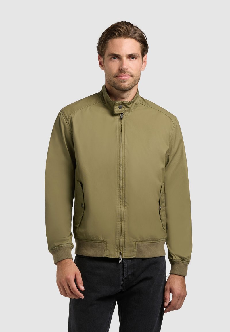 Homme portant une veste zippée vert olive avec col boutonné et poches latérales, debout devant un fond gris clair uni.