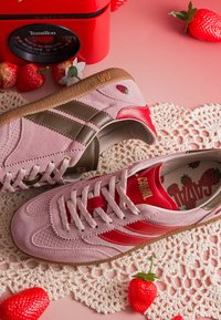 Baskets en daim roses et rouges avec des rayures marron, broderie fraise, posées sur un tapis au crochet entouré de fraises artificielles et une balance rouge.