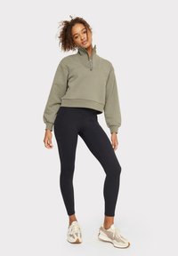 Sudadera recortada color oliva con media cremallera, mangas abullonadas, combinada con leggings negros y zapatillas multicolores. Tela suave y diseño ajustado.