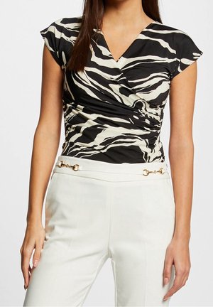 Frau trägt ein schwarz-weiß gestreiftes Wickeltop im Zebra-Design mit kurzen Ärmeln und hoch taillierte weiße Hose mit goldfarbenen Horsebit-Details.