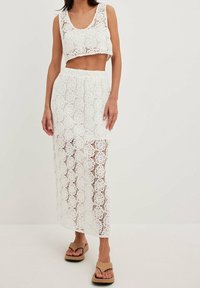 Frau, die ein weißes Häkelcroptop und einen passenden langen Rock mit beigen Flip-Flop-Sandalen trägt, steht vor einem schlichten hellen Hintergrund.