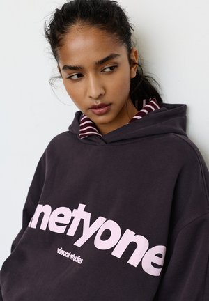 Tmavomodrá sivá mikina s veľkým svetloružovým logom s nápisom „netyone“ a „visual studies“. Obsahuje pruhovaný golier pod kapucňou. Mäkká textúra látky.