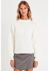Pull en tricot blanc avec un motif en diamant texturé, col rond et manches à bord côtes. Porté sur un skort à carreaux gris.