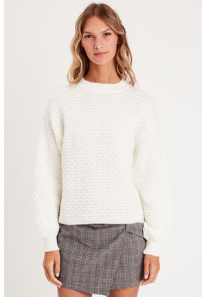 Maglione bianco lavorato a maglia con un motivo a diamante testurizzato, scollatura rotonda e polsini a costine. Indossato su una gonnellina a quadri grigia.