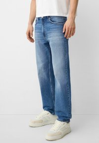 Jeans de corte relajado en azul claro, con un acabado desgastado, cinco bolsillos y cierre de botón, combinados con zapatillas blancas.