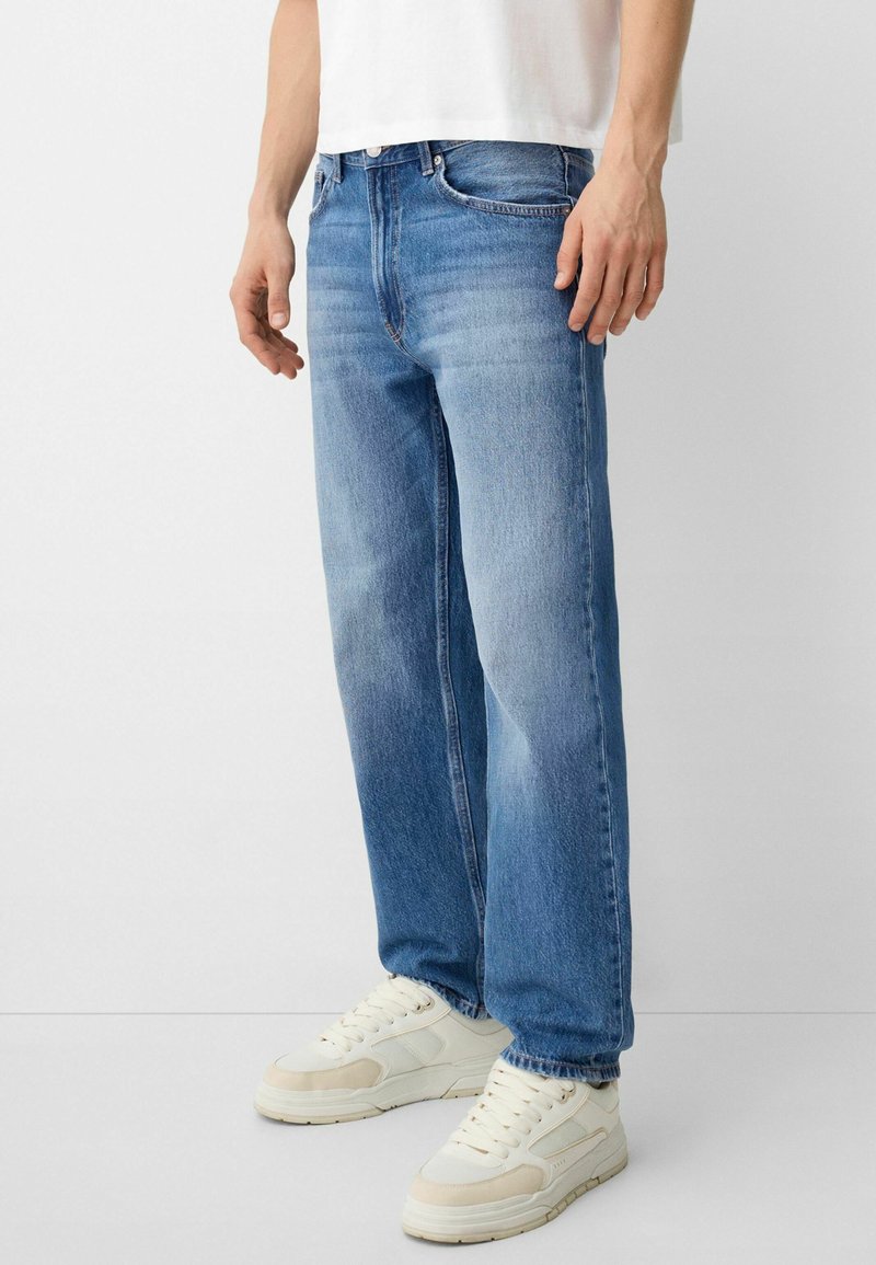 Jeans de corte relajado en azul claro, con un acabado desgastado, cinco bolsillos y cierre de botón, combinados con zapatillas blancas.