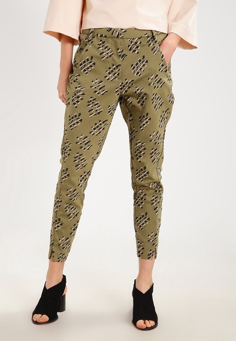 Personne portant un pantalon à motif vert olive, un haut clair à manches larges et des chaussures à talons noires ouvertes aux orteils, debout les mains dans les poches.