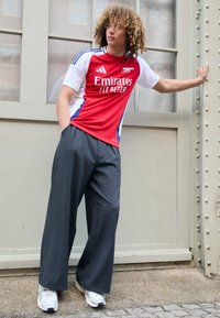 Tricou scurt cu mânecă roșie și albă, cu accente albastre, combinat cu pantaloni wide-leg gri și adidași albi, sprijinindu-se de un perete.