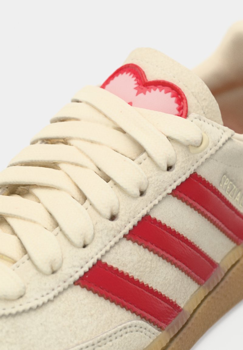Leather retro sneakers with red stripes, beige upper, and textured tongue featuring a pink heart detail. Gum sole provides contrast.