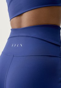 Niebieskie legginsy z wysokim stanem, wykonane z gładkiego, elastycznego materiału, z wygiętym szwem i subtelnym logo z tyłu.