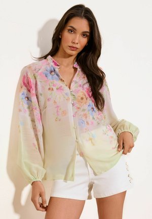 Vrouw met lang donker haar draagt een pastelgekleurde, gebloemde blouse met knopen en opgeblazen mouwen en witte shorts tegen een effen lichte achtergrond.