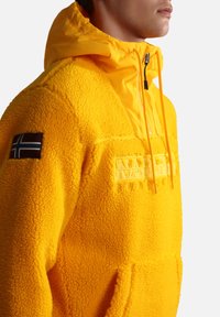 Yellow fleece pullover with a textured surface, a hood, and front pocket. Features embroidered "NAPAPIJRI" logo and an insignia patch on the sleeve.