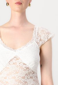 Lace topp i hvitt med korte puffermer, med en bølgete halslinje og blomstermønster. Silkeaktig tekstur med gjennomsiktige elementer.