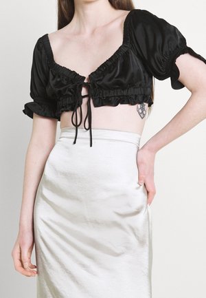 Mujer vistiendo un top corto negro arrugado con mangas abullonadas y una falda blanca de satén de talle alto, con la mano apoyada en la cadera, tatuaje visible.