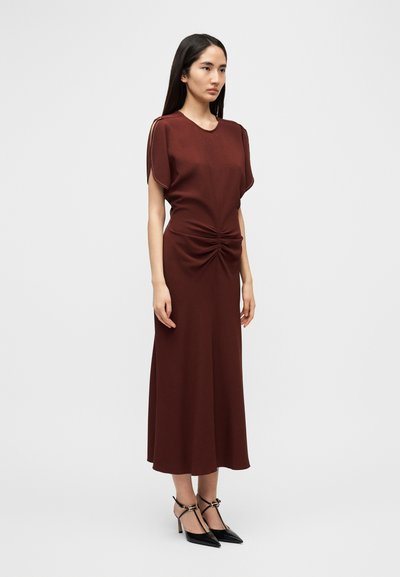 Μπορντό, φόρεμα μέχρι το γόνατο με κοντά μανίκια και σούρες στη μέση· συνδυασμένο με μαύρα ψηλοτάκουνα παπούτσια με μυτερή μύτη. Ομαλή υφή υφάσματος.