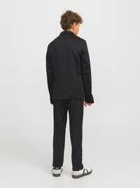 Czarny garnitur, składający się z marynarki i spodni o gładkiej fakturze. Marynarka ma klasyczny krój, a obuwie jest białe z czarnymi akcentami.