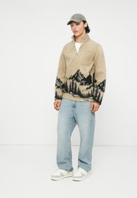 Beiger Fleece-Pullover mit einem Viertelreißverschluss und schwarzem Berglandschaftsdesign, kombiniert mit hellblauen Jeans und weißen Schuhen.