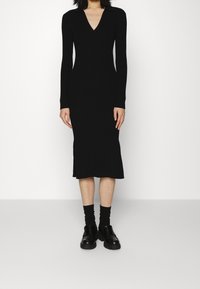 Abito nero a costine con scollo a V e maniche lunghe, caratterizzato da una silhouette aderente e spacchi laterali. Abbinato a scarpe con plateau nere e calzini.