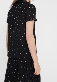 Blusa nera con maniche corte a volant, decorata con motivi di stelle dorate. Realizzata in tessuto leggero, presenta una texture liscia e un design semplice.