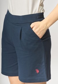 Pantaloni corti blu navy realizzati in tessuto morbido, con vita elasticizzata, tasche laterali e un piccolo logo ricamato rosso sulla coscia sinistra.
