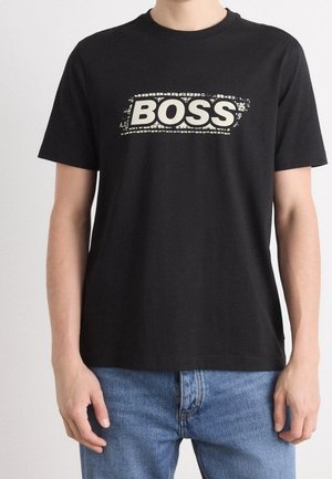 Personne portant un t-shirt noir avec un texte blanc usé "BOSS" sur la poitrine, associé à un jean bleu, debout devant un fond uni.