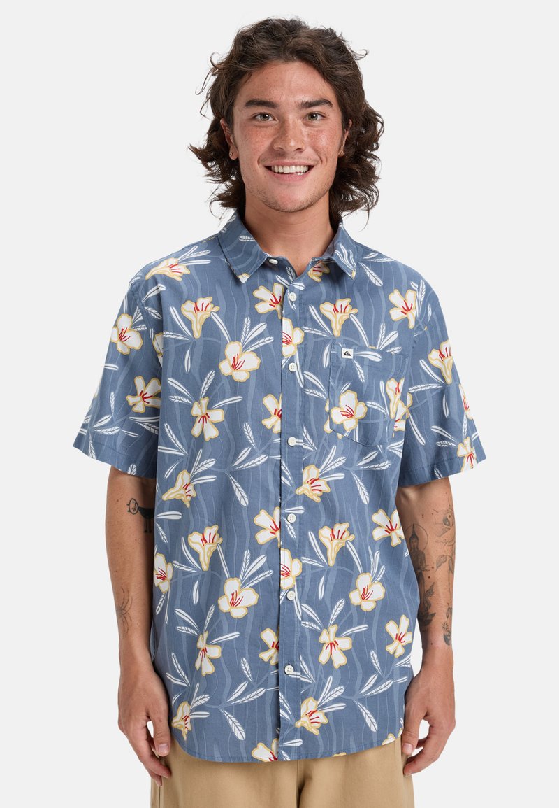 Košile na knoflíky s krátkým rukávem v modré barvě s bílým a žlutým květinovým vzorem. Obsahuje kapsu a lehkou texturovanou tkaninu.