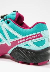 Türkis- und magentafarbener Laufschuh mit strukturiertem Mesh-Obermaterial, markantem Seitendesign und auffälliger Markenkennzeichnung; ausgestattet mit einer schwarzen Gummisohle.
