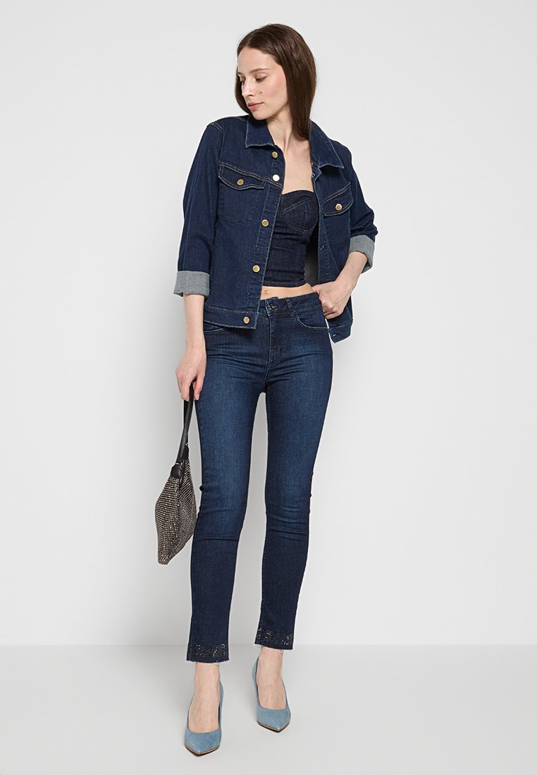 Giacca di jeans con bottoni dorati, maniche arrotolate; top corto in denim blu scuro; jeans slim fit blu scuro; orlo accentuato; tacchi blu; borsa in rete.