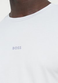 Valge puuvillane t-särk, millel on ümar kaelus ja ees väike lilla "BOSS" logo, mis näitab siledat tekstuuri.