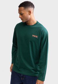 Giovane uomo che guarda verso il basso, indossa una maglia a maniche lunghe verde scuro con piccoli testi rossi e bianchi sul petto a sinistra, abbinata a jeans blu.