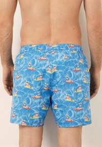 Un maillot de bain bleu ornés d'un motif de chiens surfant sur des planches colorées avec des vagues, fabriqué en tissu léger avec une taille élastique.
